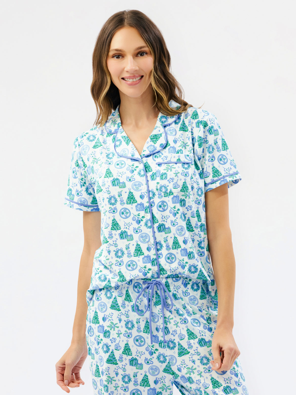 A woman modeling a matching short-sleeve pajama top and shorts set with a white base and a blue and green holiday tree pattern, set against a plain white background.