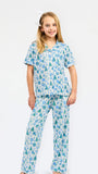A young girl modeling a two-piece pajama set with a festive holiday print of pine trees, ornaments, and snowflakes in shades of blue and green on a white background, set against a plain white studio backdrop.