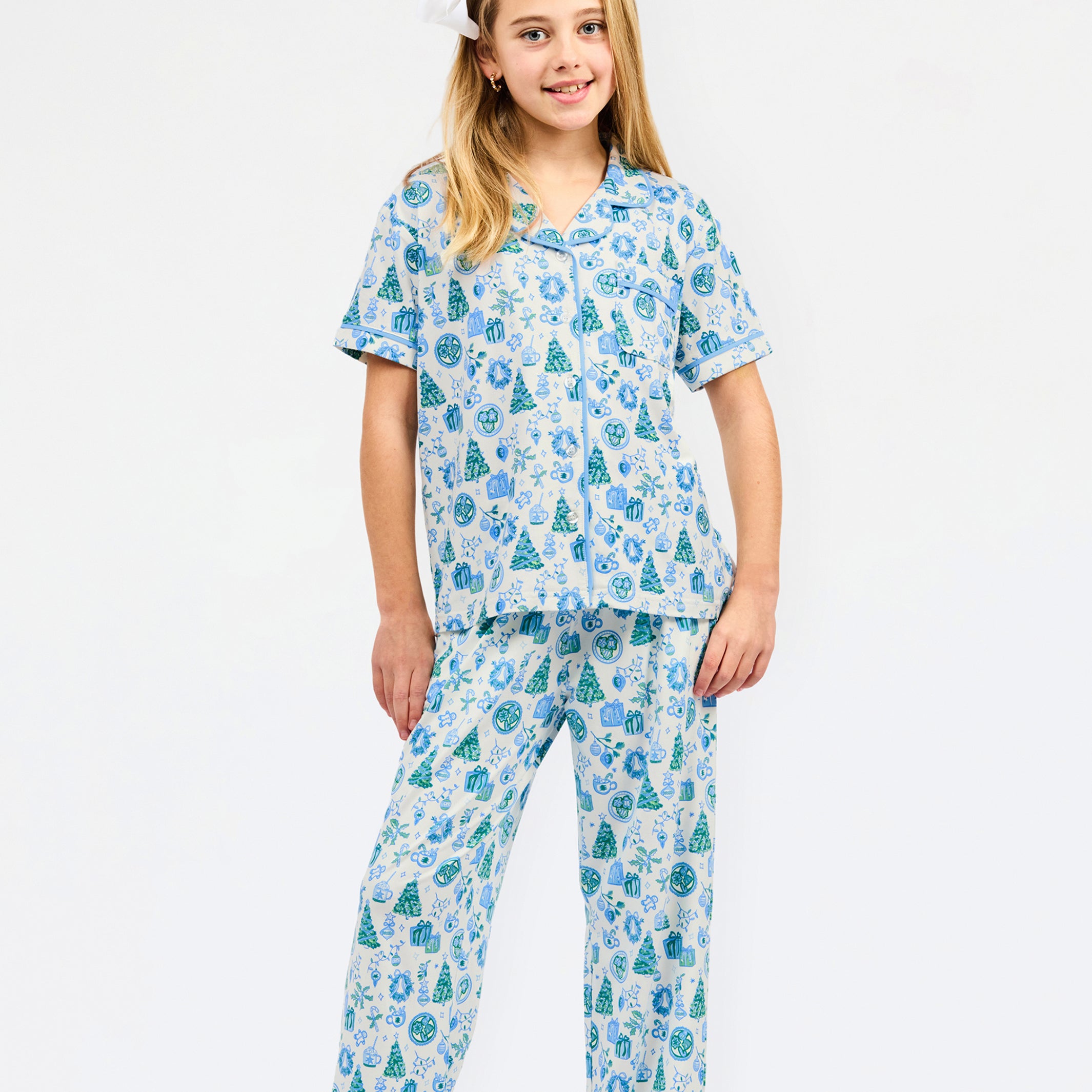 A young girl modeling a two-piece pajama set with a festive holiday print of pine trees, ornaments, and snowflakes in shades of blue and green on a white background, set against a plain white studio backdrop.