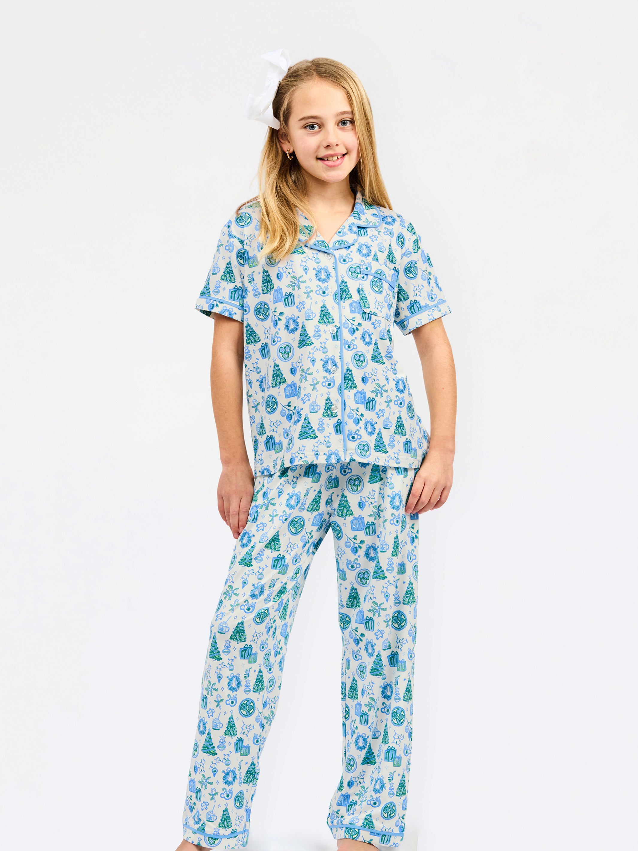 A young girl modeling a two-piece pajama set with a festive holiday print of pine trees, ornaments, and snowflakes in shades of blue and green on a white background, set against a plain white studio backdrop.