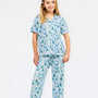 A young girl modeling a two-piece pajama set with a festive holiday print of pine trees, ornaments, and snowflakes in shades of blue and green on a white background, set against a plain white studio backdrop.