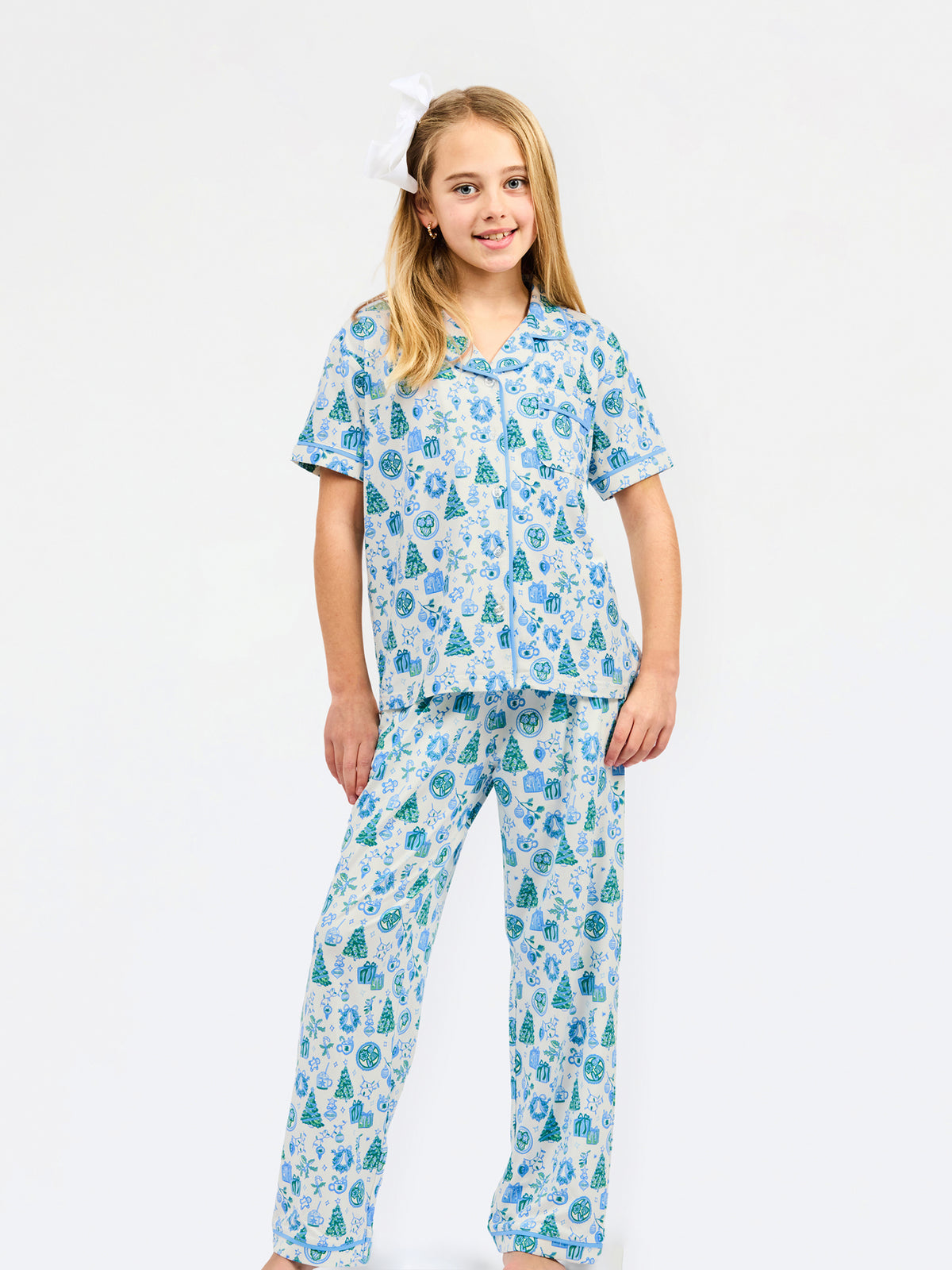 A young girl modeling a two-piece pajama set with a festive holiday print of pine trees, ornaments, and snowflakes in shades of blue and green on a white background, set against a plain white studio backdrop.