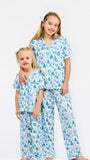 Two young girls wearing matching pajama sets with a blue and green Christmas tree pattern on a white background, standing against a plain white studio backdrop.