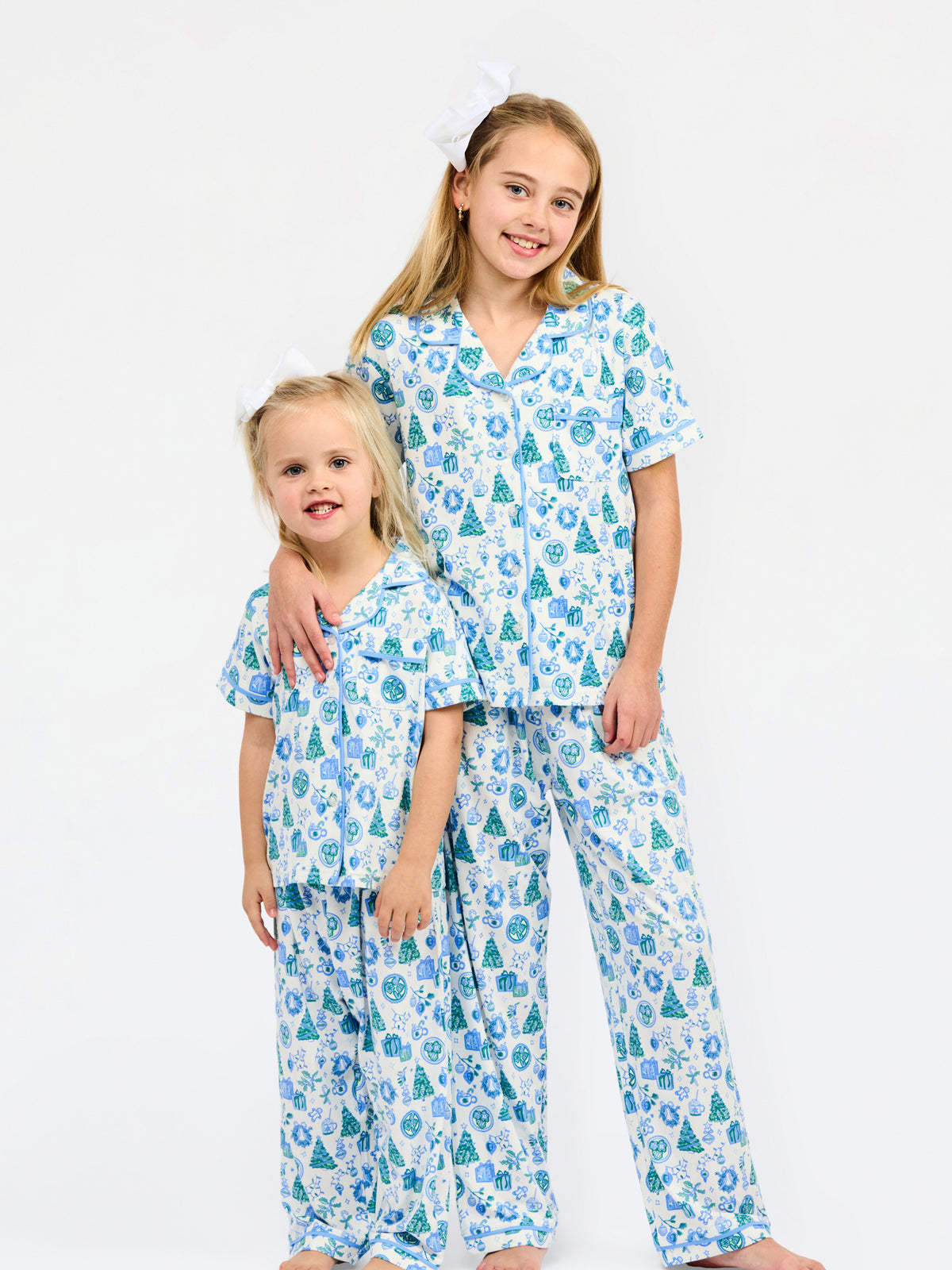 Two young girls wearing matching pajama sets with a blue and green Christmas tree pattern on a white background, standing against a plain white studio backdrop.