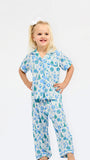 A young girl with blonde hair tied up with a white bow is wearing a two-piece pajama set with a blue and green Christmas tree pattern on a white background, standing against a plain white studio background.