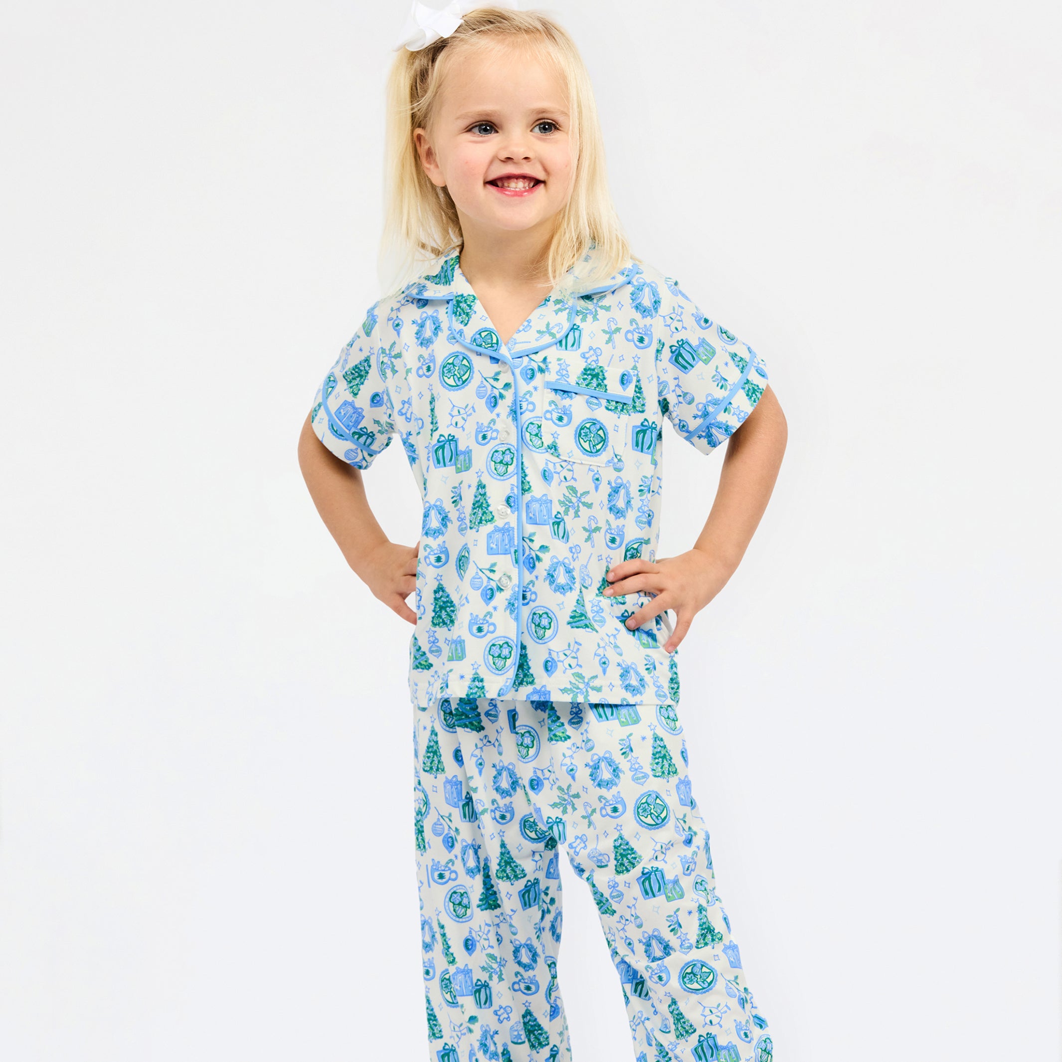 A young girl with blonde hair tied up with a white bow is wearing a two-piece pajama set with a blue and green Christmas tree pattern on a white background, standing against a plain white studio background.