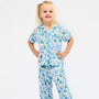 A young girl with blonde hair tied up with a white bow is wearing a two-piece pajama set with a blue and green Christmas tree pattern on a white background, standing against a plain white studio background.