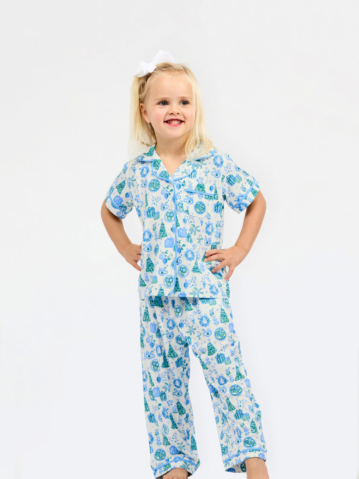 A young girl with blonde hair tied up with a white bow is wearing a two-piece pajama set with a blue and green Christmas tree pattern on a white background, standing against a plain white studio background.