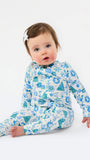 A baby sitting on a white studio background wearing a long-sleeve footed sleeper with a blue and green winter-themed pattern of snow globes, trees, and snowflakes.