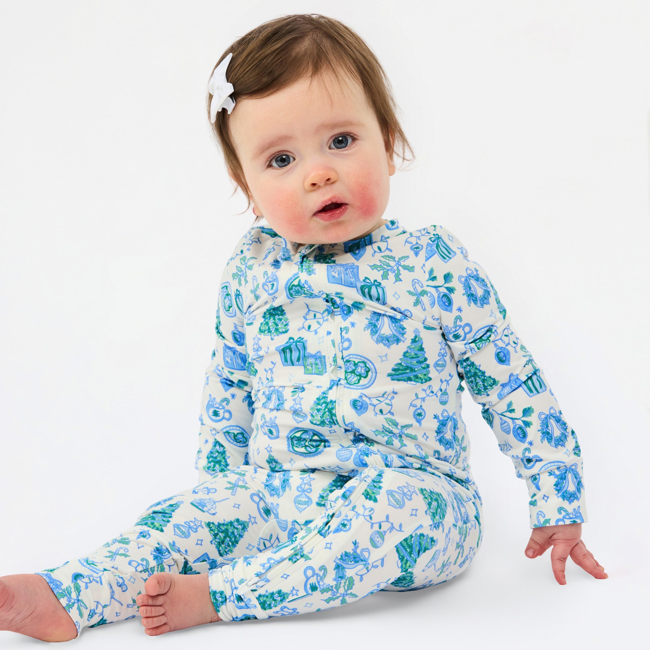 A baby sitting on a white studio background wearing a long-sleeve footed sleeper with a blue and green winter-themed pattern of snow globes, trees, and snowflakes.