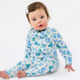 A baby sitting on a white studio background wearing a long-sleeve footed sleeper with a blue and green winter-themed pattern of snow globes, trees, and snowflakes.