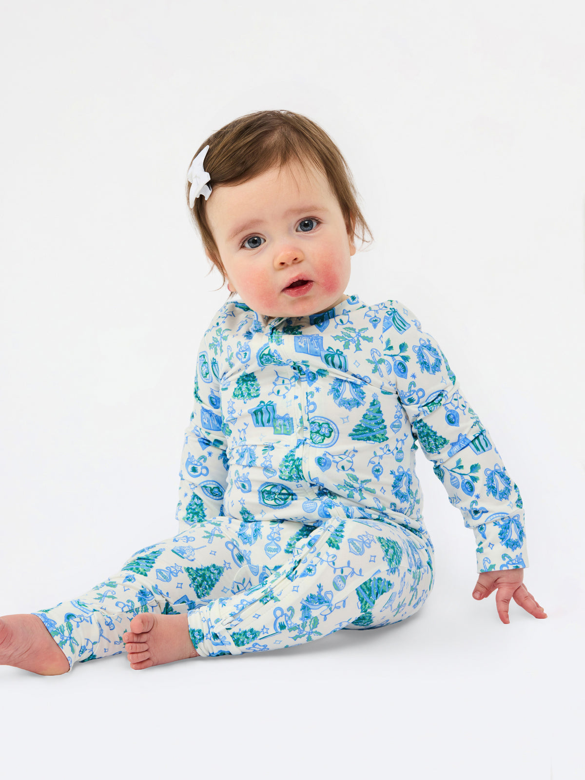 A baby sitting on a white studio background wearing a long-sleeve footed sleeper with a blue and green winter-themed pattern of snow globes, trees, and snowflakes.