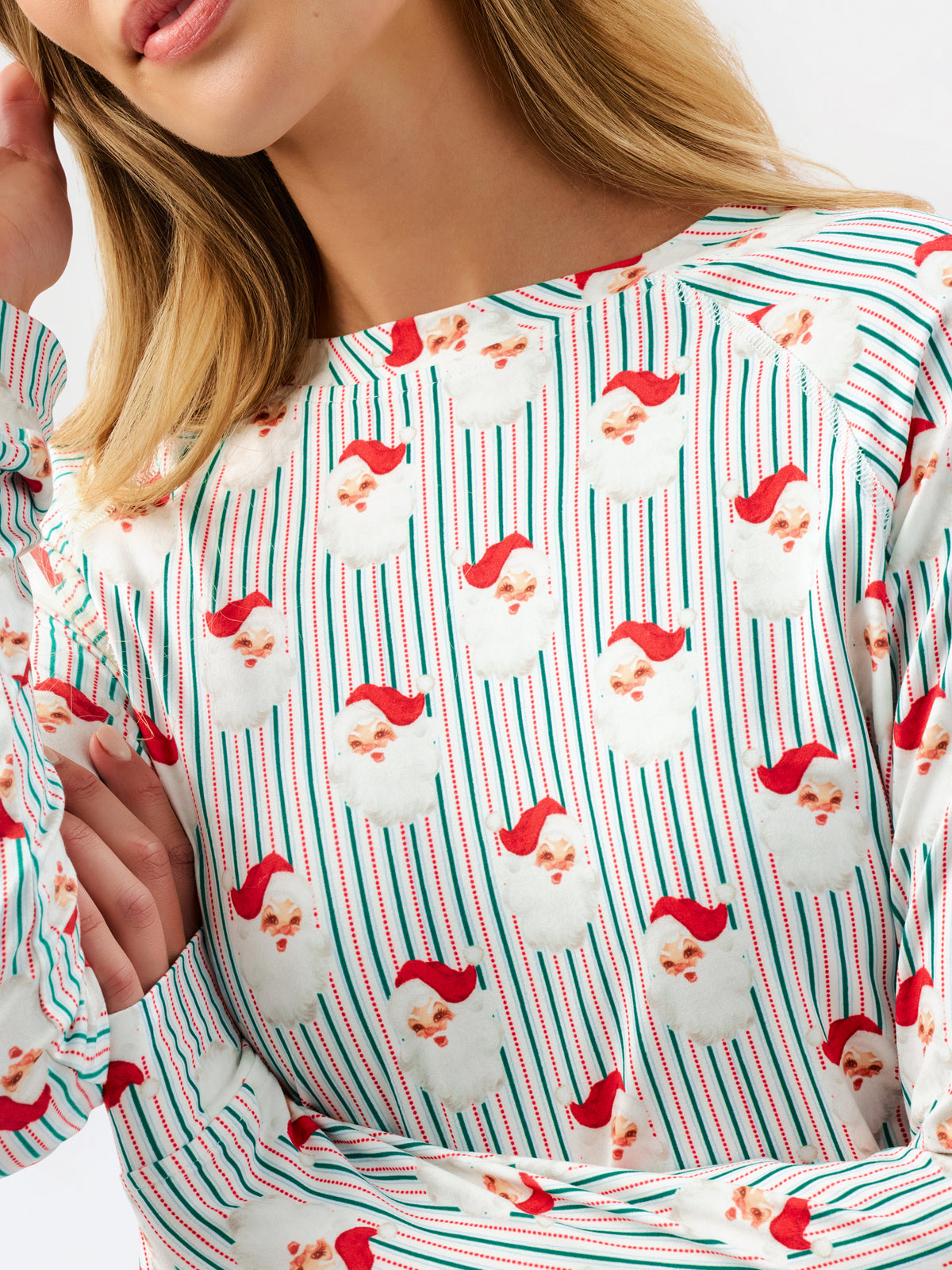 A close-up of a woman wearing a long-sleeve top with a festive holiday print of Santa Claus faces over a white background with thin red and green vertical stripes.