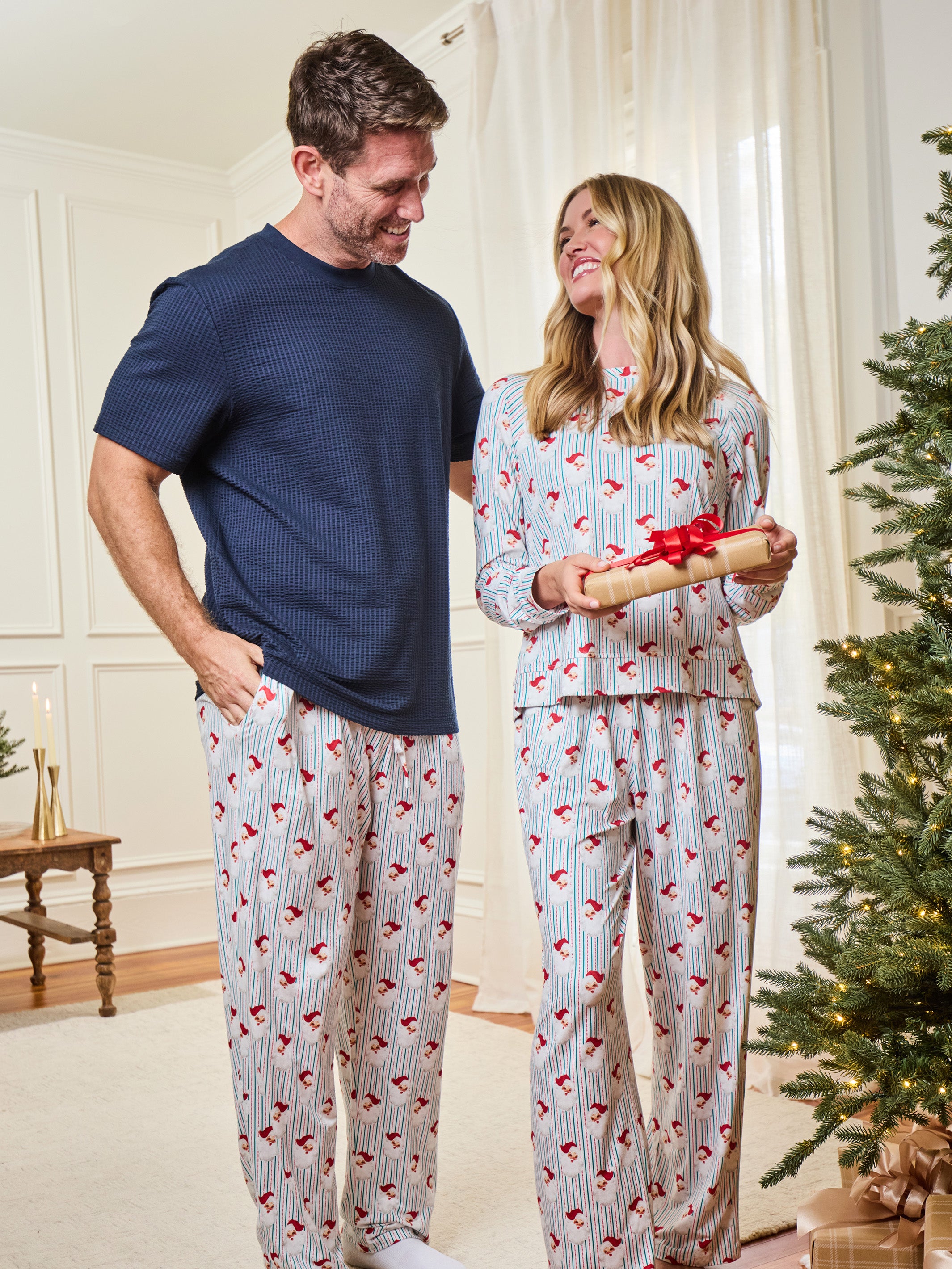 A man and a woman wearing matching holiday-themed waffle-knit pajamas stand in a living room next to a Christmas tree. The man wears a navy blue t-shirt with white pinstriped pajama pants featuring a red heart pattern.