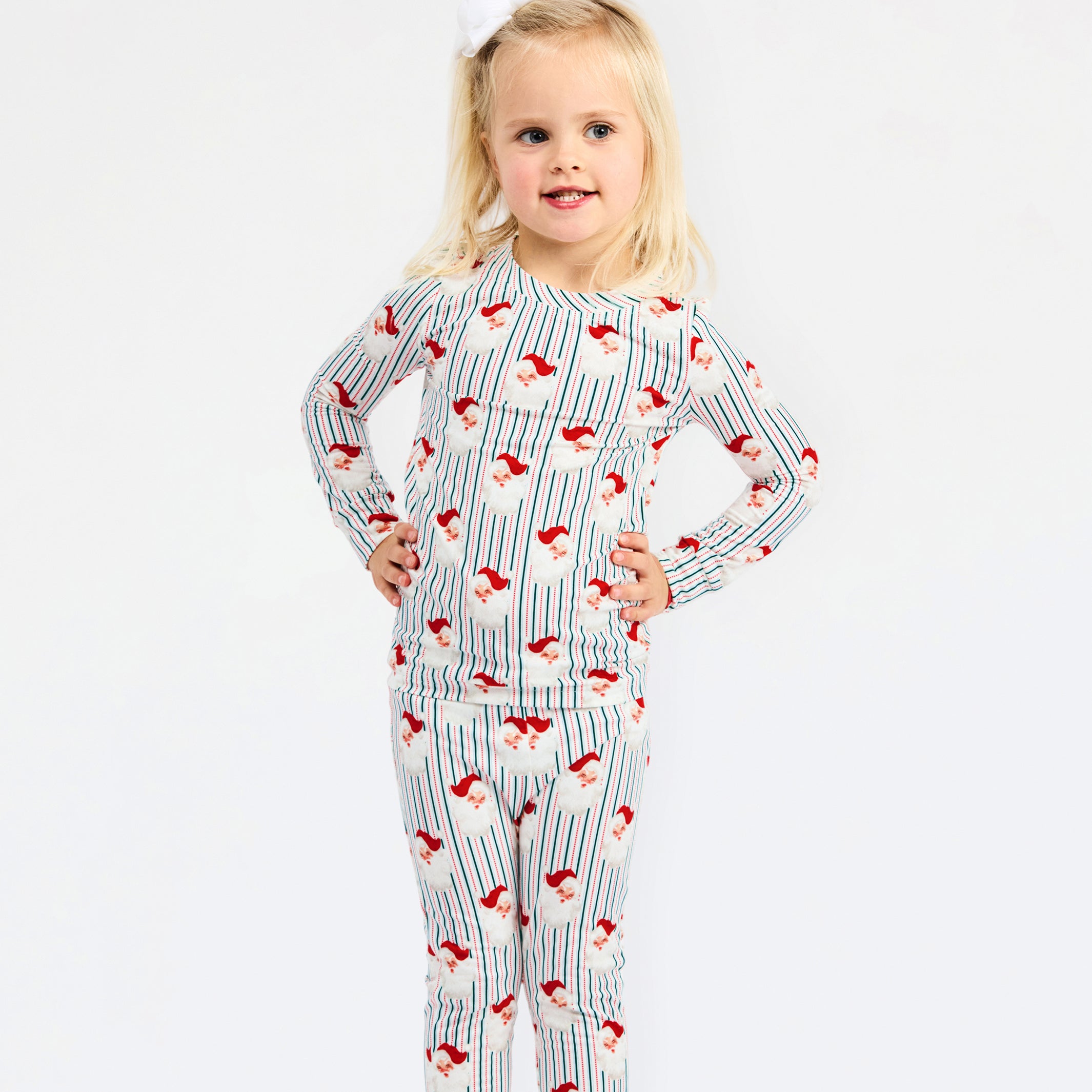 A young blonde girl wearing a long-sleeve pajama set with a blue and white pinstripe pattern and red Santa Claus motifs, standing against a plain white background.