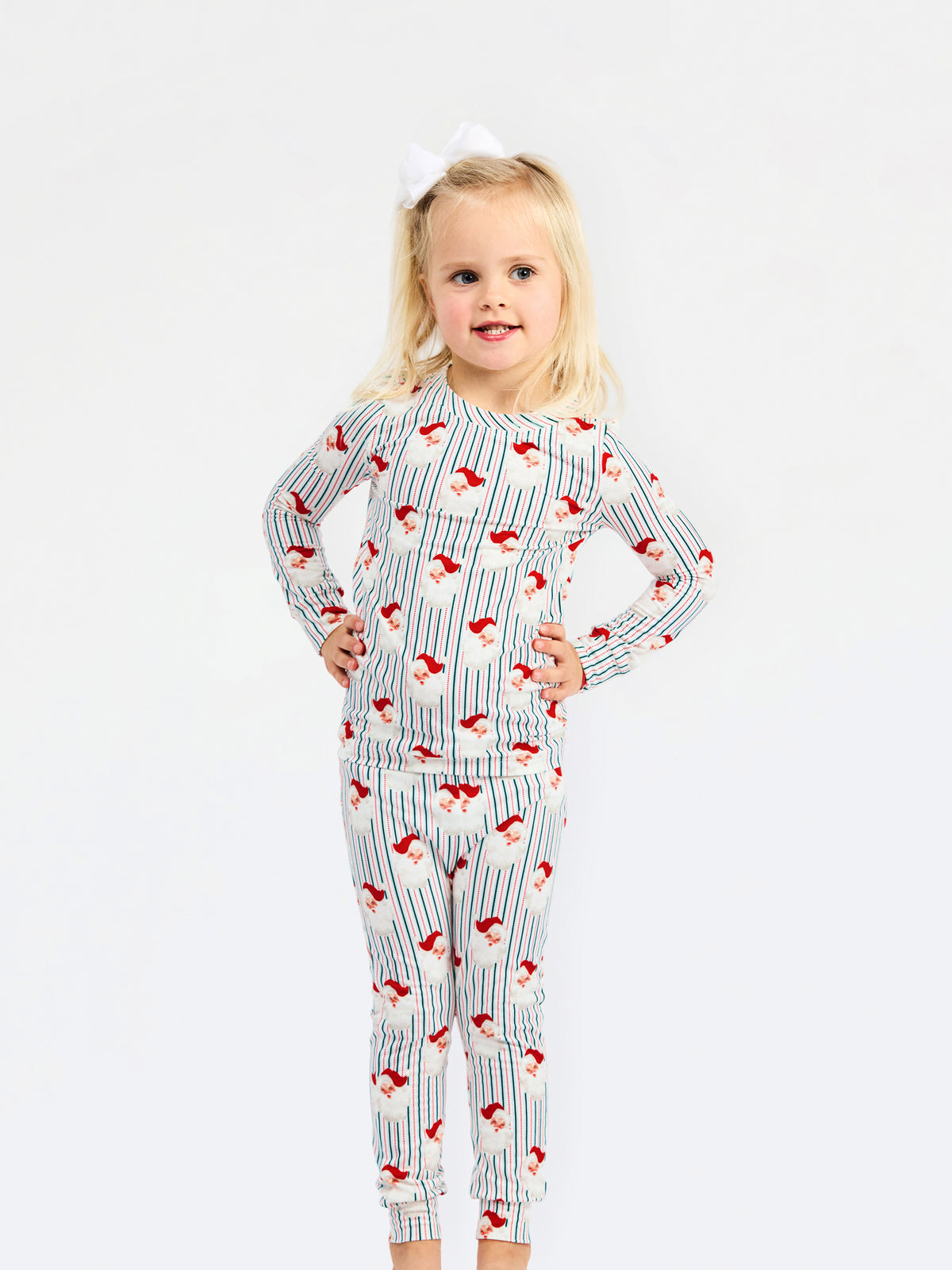 A young blonde girl wearing a long-sleeve pajama set with a blue and white pinstripe pattern and red Santa Claus motifs, standing against a plain white background.