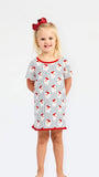 A young girl with blonde hair and a red bow stands against a white studio background wearing a short-sleeve nightgown with a white base, red trim, and a blue and red pinstripe pattern.