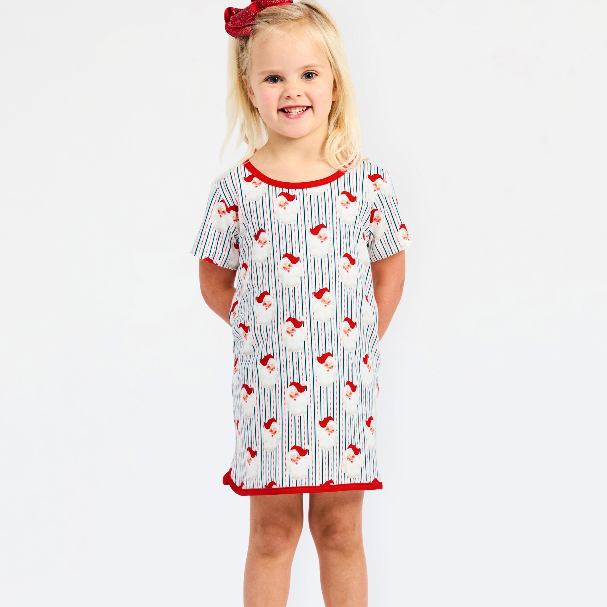 A young girl with blonde hair and a red bow stands against a white studio background wearing a short-sleeve nightgown with a white base, red trim, and a blue and red pinstripe pattern.