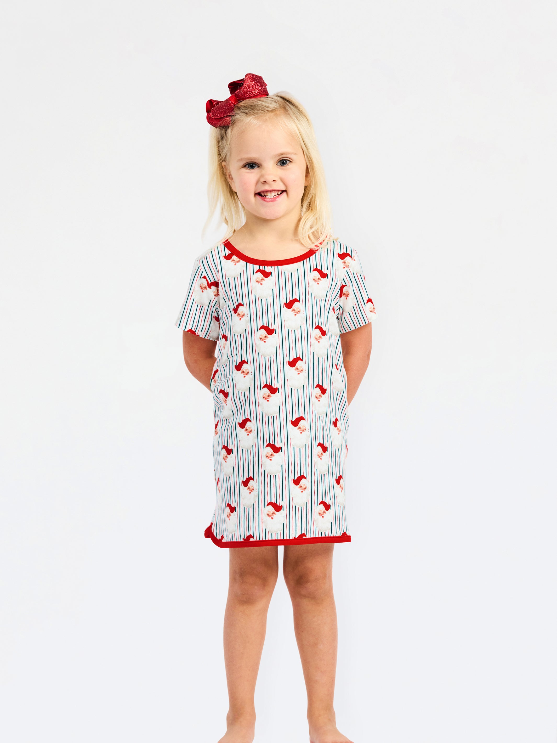 A young girl with blonde hair and a red bow stands against a white studio background wearing a short-sleeve nightgown with a white base, red trim, and a blue and red pinstripe pattern.