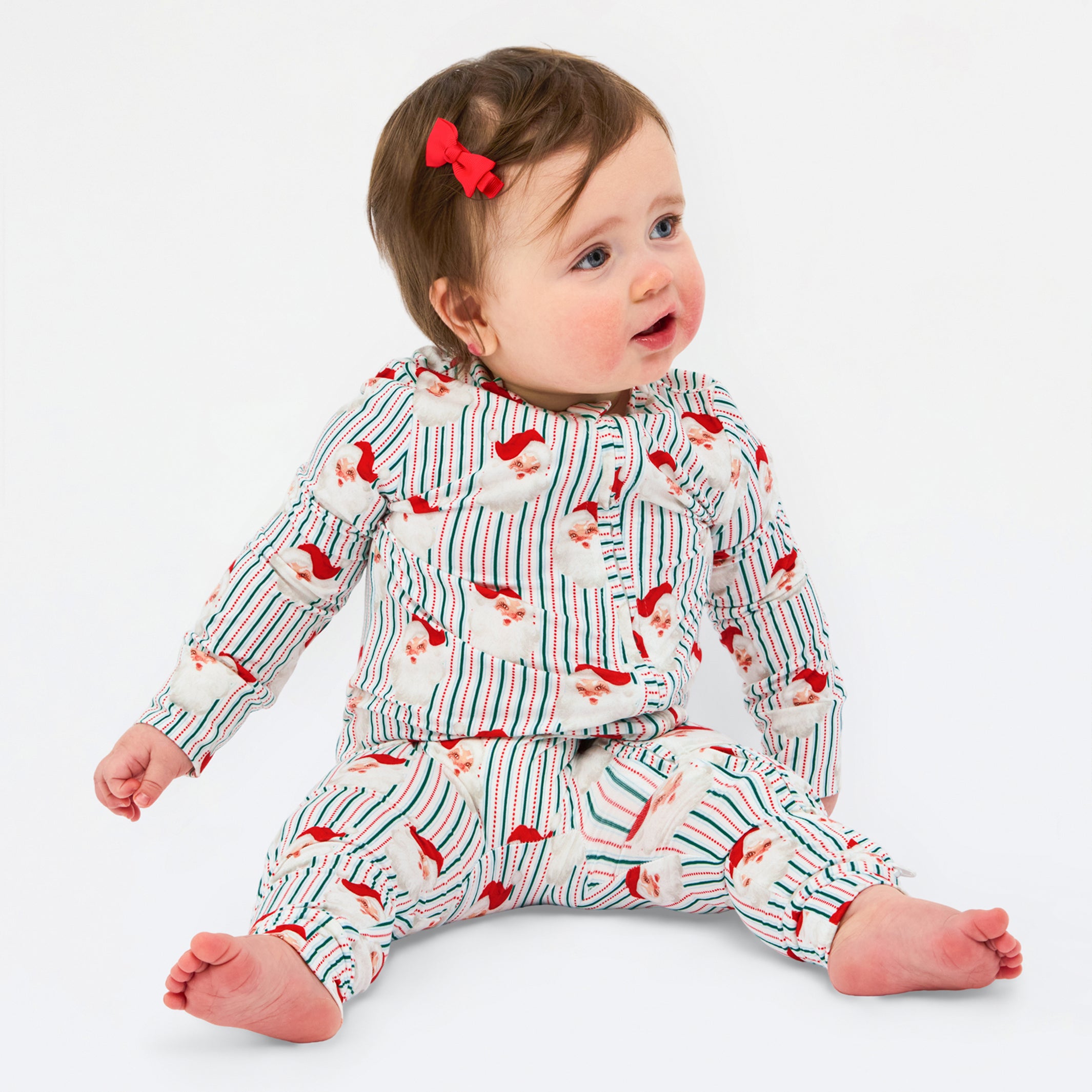 A baby sitting on a white studio background wearing a matching long-sleeve top and pants set with a red and blue striped pattern and small red bird motifs. The baby has a small red bow in their hair.