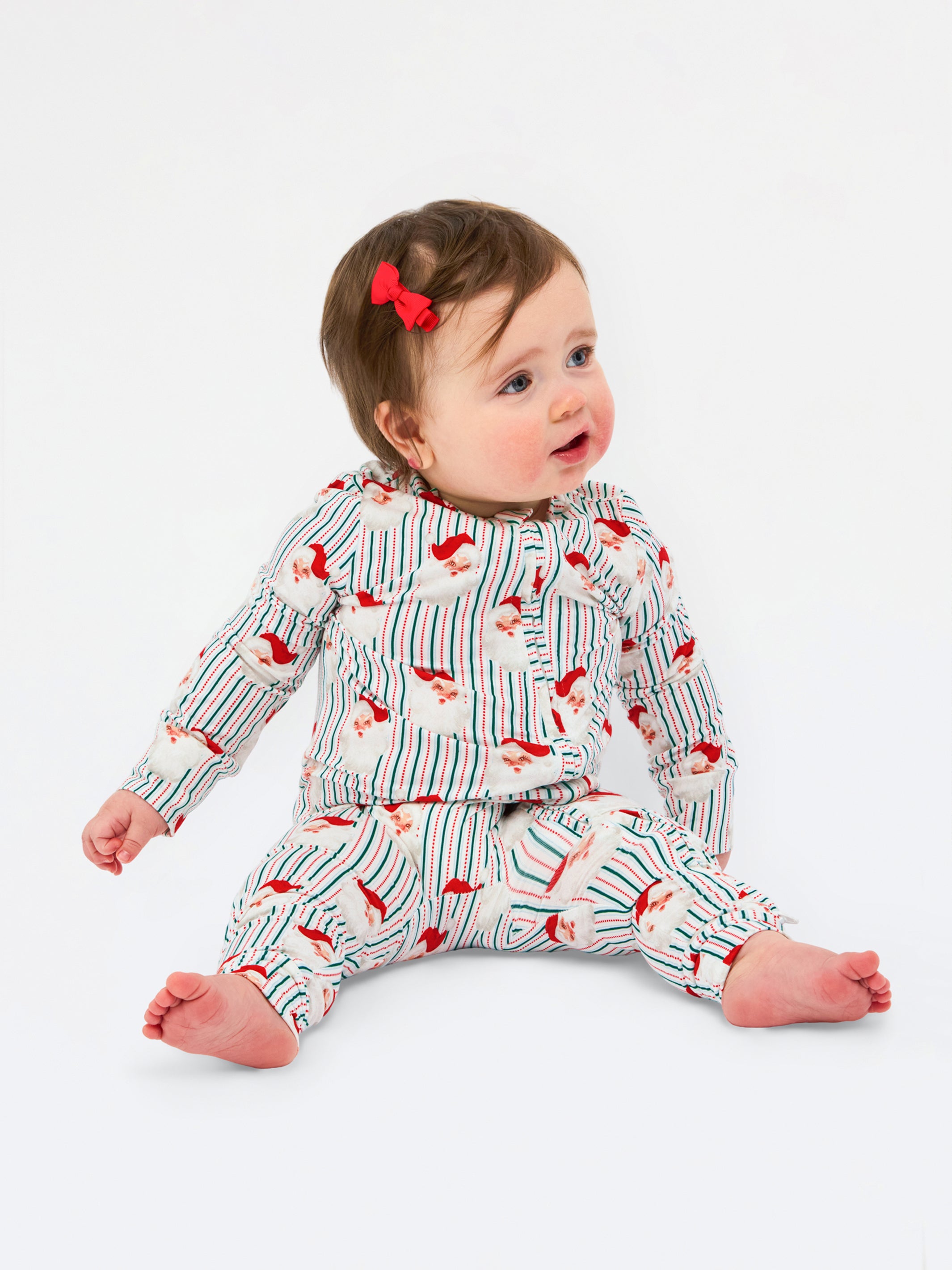 A baby sitting on a white studio background wearing a matching long-sleeve top and pants set with a red and blue striped pattern and small red bird motifs. The baby has a small red bow in their hair.