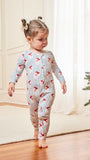 A toddler girl walking barefoot indoors wearing a long-sleeve footless pajama romper with thin blue vertical stripes and red cartoon horse motifs, next to a decorated Christmas tree and wrapped gift box.