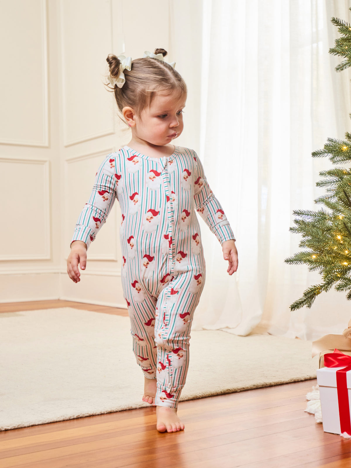 A toddler girl walking barefoot indoors wearing a long-sleeve footless pajama romper with thin blue vertical stripes and red cartoon horse motifs, next to a decorated Christmas tree and wrapped gift box.