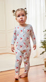 A toddler stands barefoot on a wooden floor indoors, wearing a long-sleeve footless pajama romper with thin blue vertical stripes and a red holiday print. A Christmas tree and wrapped gift are visible in the softly lit room.