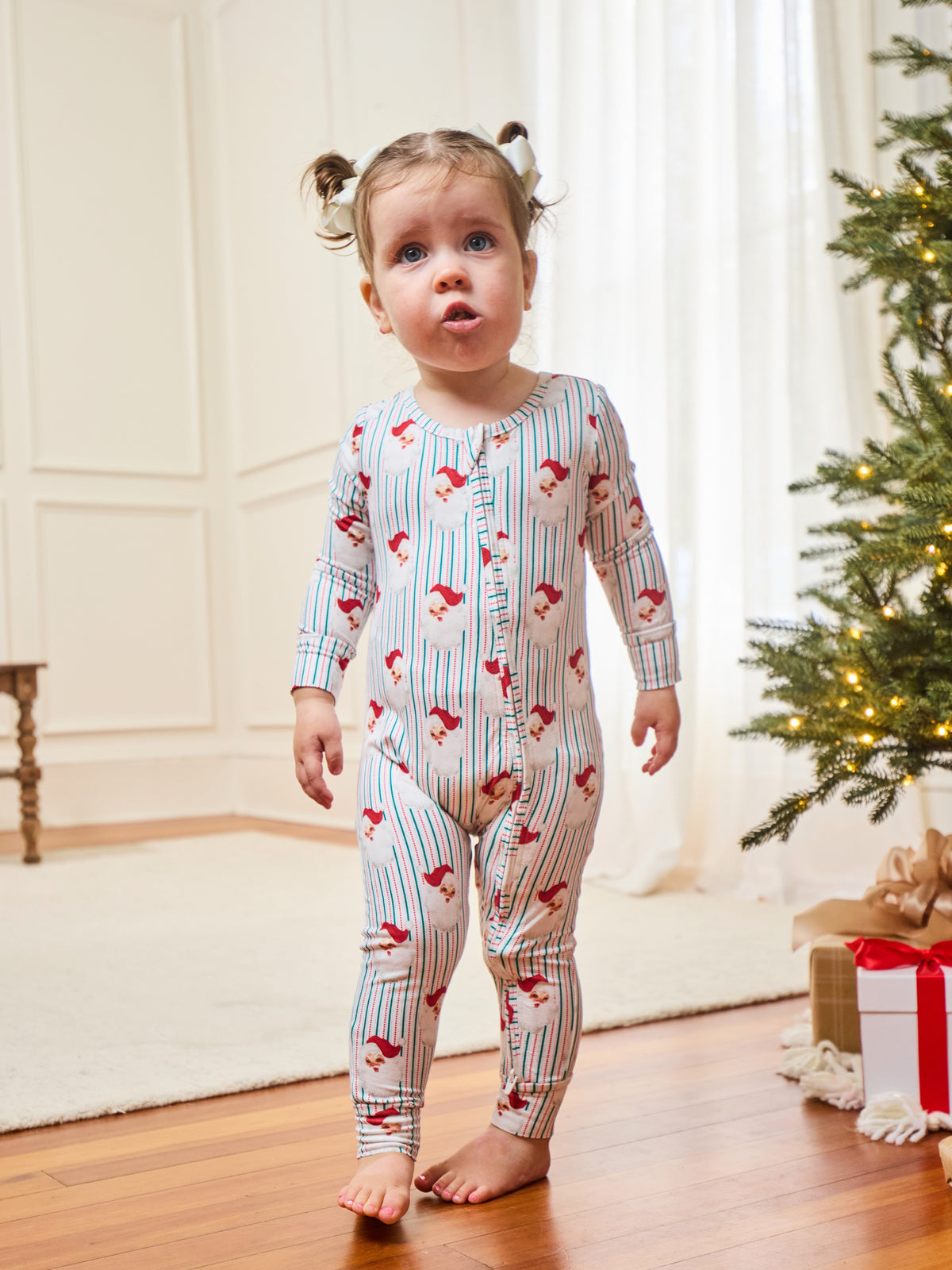 A toddler stands barefoot on a wooden floor indoors, wearing a long-sleeve footless pajama romper with thin blue vertical stripes and a red holiday print. A Christmas tree and wrapped gift are visible in the softly lit room.
