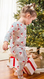 A toddler wearing a festive Christmas-themed long-sleeve pajama set with red Santa prints and blue vertical stripes, standing near wrapped gifts under a decorated Christmas tree.