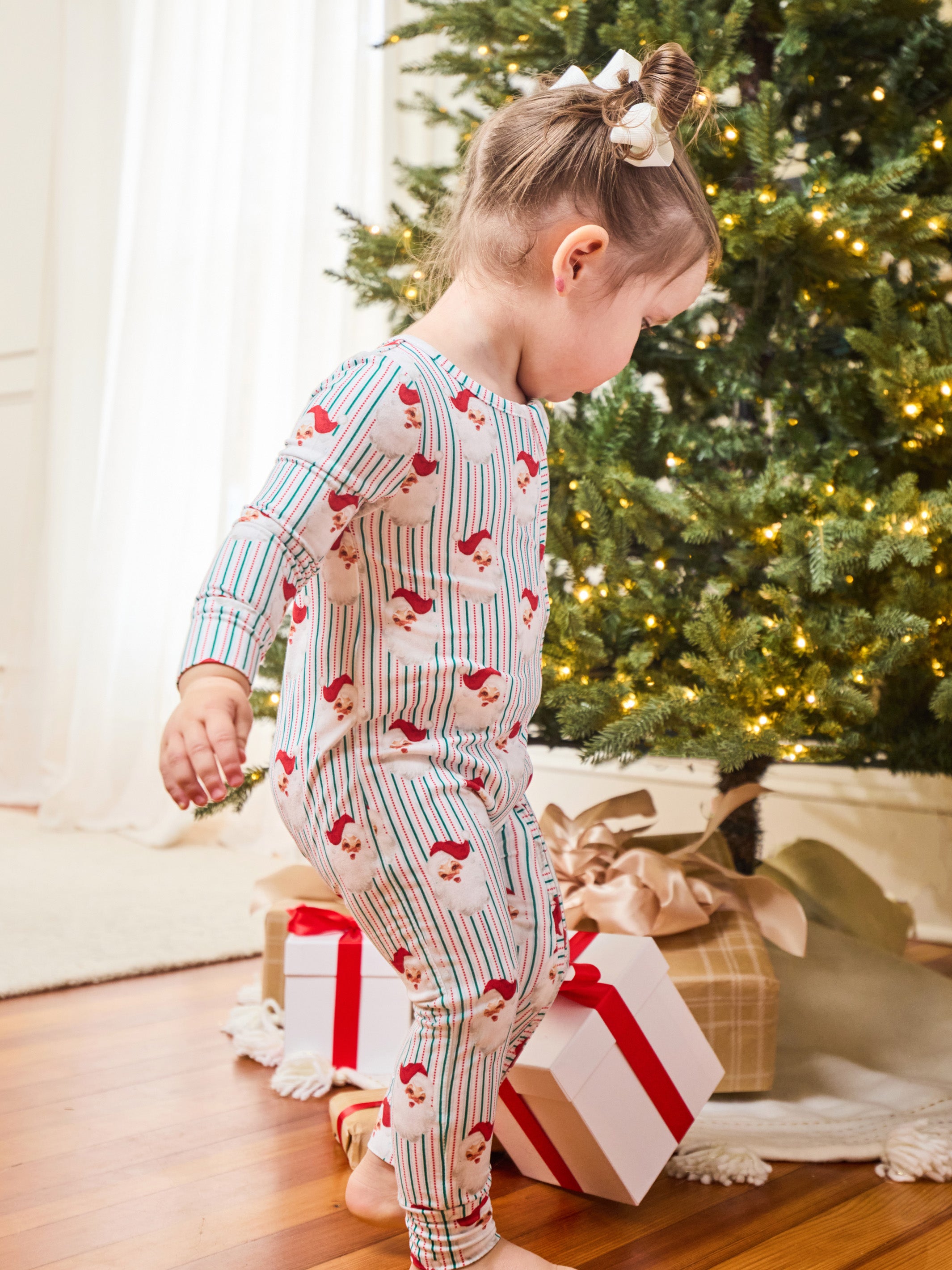 A toddler wearing a festive Christmas-themed long-sleeve pajama set with red Santa prints and blue vertical stripes, standing near wrapped gifts under a decorated Christmas tree.