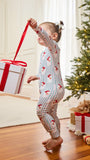 A toddler stands barefoot on a wooden floor in a living room, holding a large white gift box with a red ribbon by its loop. The child is wearing a one-piece pajama with thin red and blue vertical stripes and small red holiday motifs.
