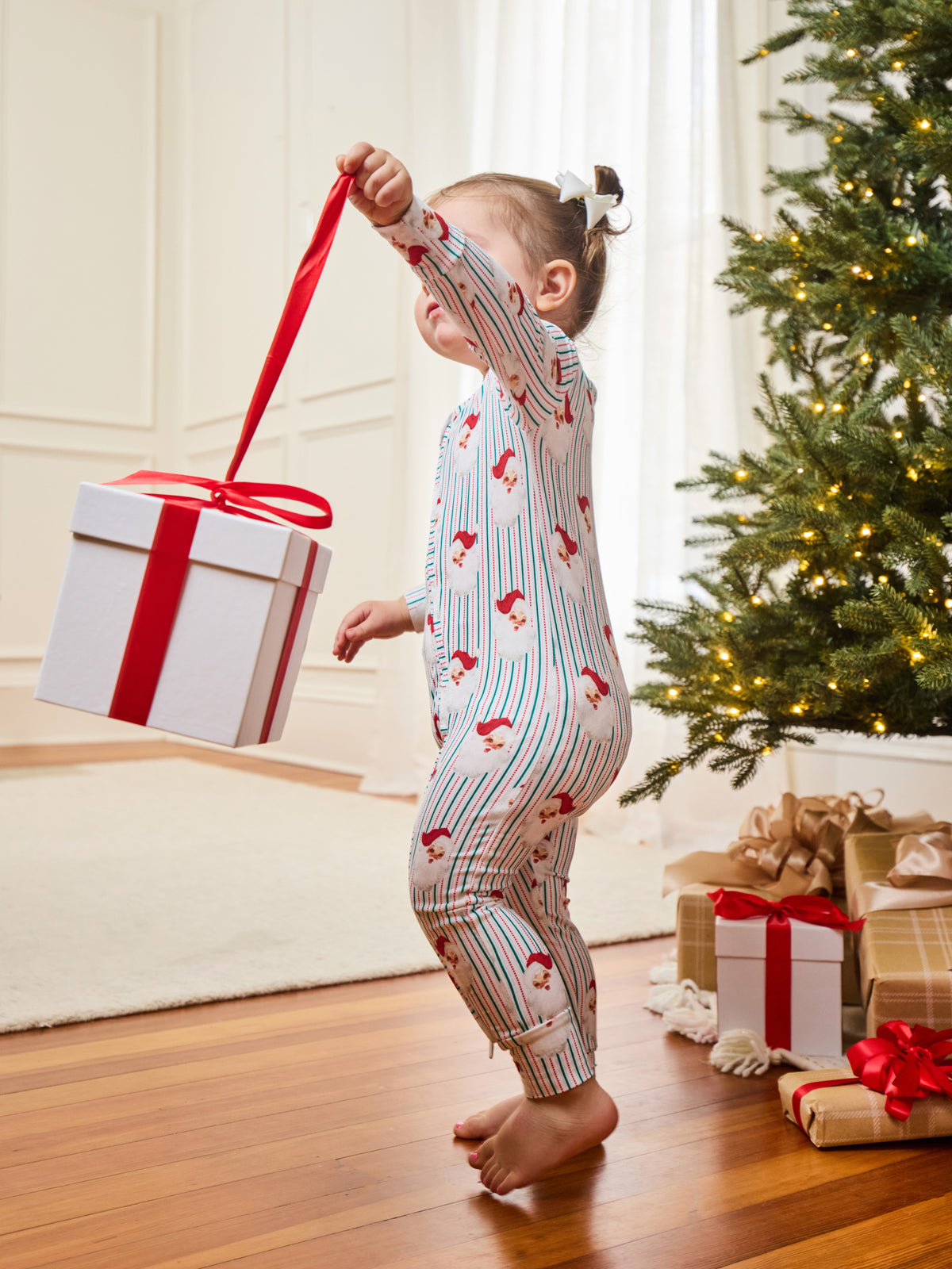 A toddler stands barefoot on a wooden floor in a living room, holding a large white gift box with a red ribbon by its loop. The child is wearing a one-piece pajama with thin red and blue vertical stripes and small red holiday motifs.
