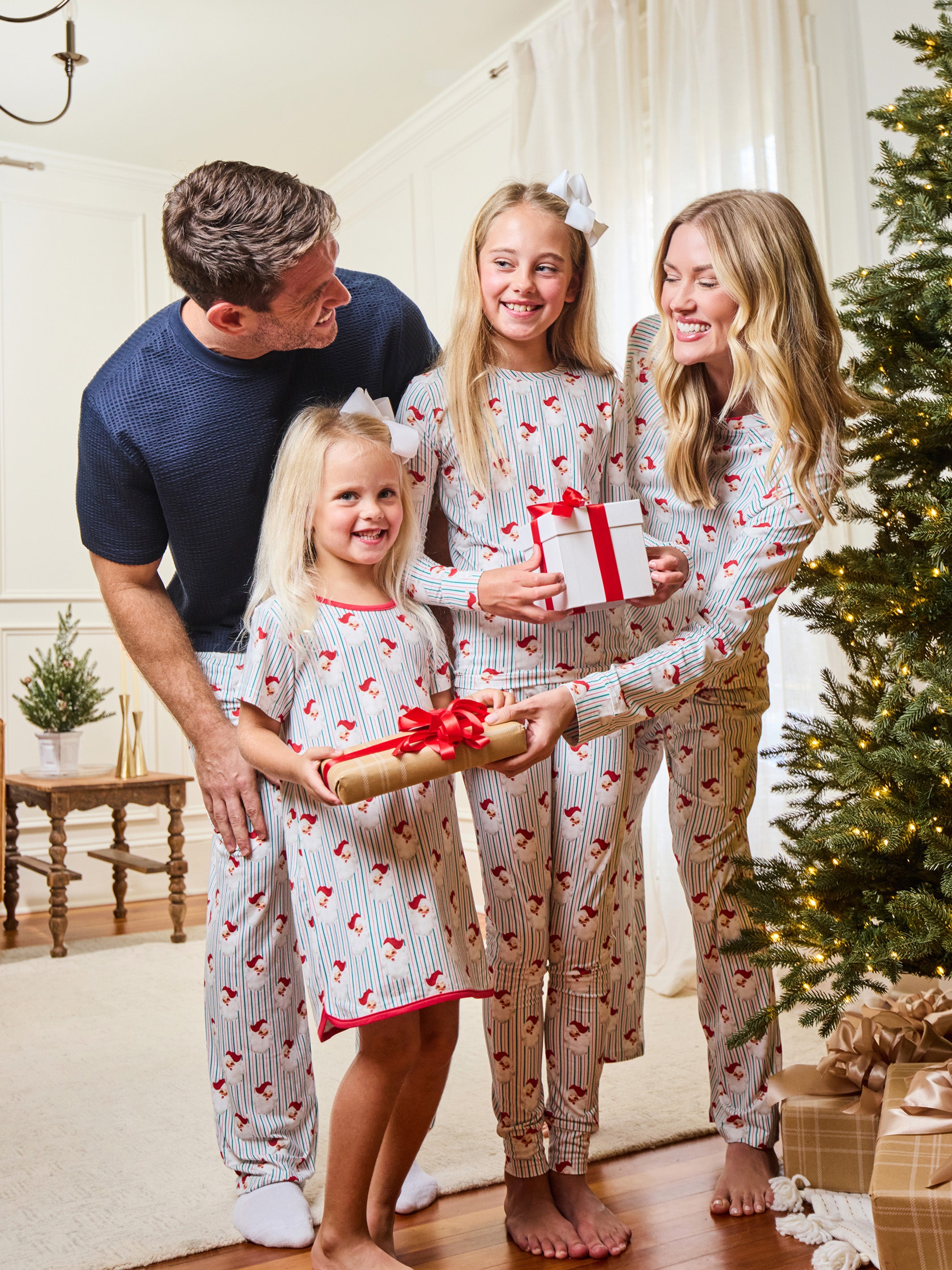 A family of four wearing matching holiday pajamas with a red heart pattern on a white background, gathered in a living room next to a Christmas tree and wrapped gifts.