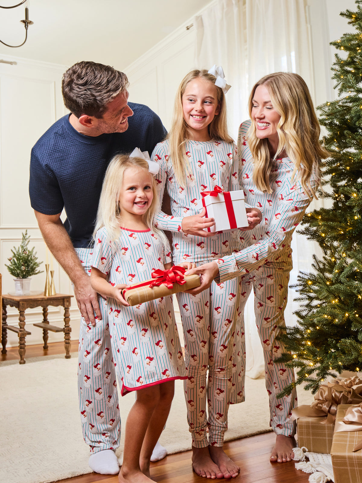 A family of four wearing matching holiday pajamas with a red heart pattern on a white background, gathered in a living room next to a Christmas tree and wrapped gifts.