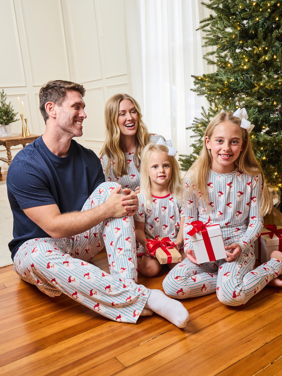 A family of four wearing matching Christmas pajamas sits together on a wooden floor in front of a decorated Christmas tree. The pajamas are white with thin red pinstripes and a red holiday pattern, and one child holds a wrapped gift with a red bow.