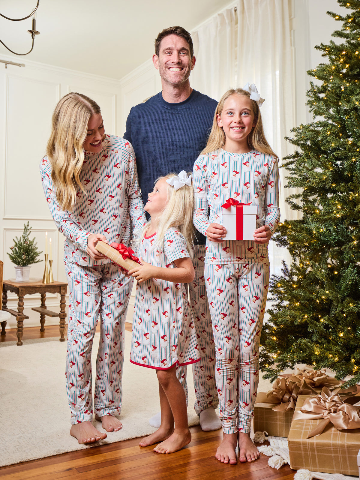 A family of four wearing matching holiday-themed pajamas stands in a living room next to a decorated Christmas tree. The pajamas are light gray with thin red stripes and a red heart pattern, with one girl holding a wrapped gift.