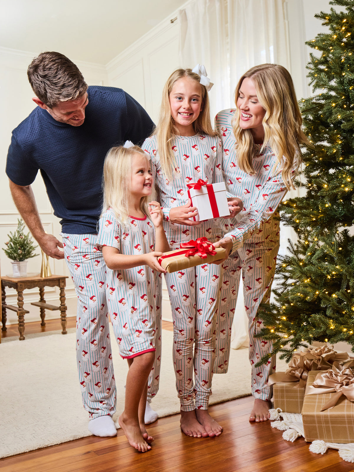 A family of four wearing matching heart-patterned pajamas stands together in a living room next to a decorated Christmas tree, holding wrapped gifts.