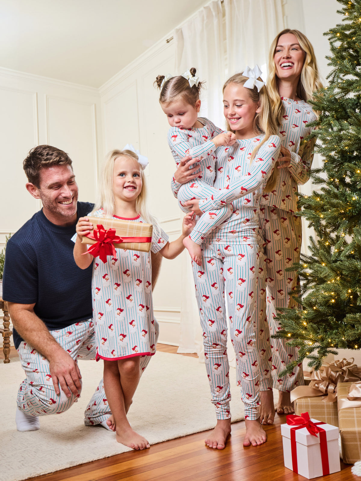 A family of five wearing matching holiday-themed pajamas poses together in a living room next to a Christmas tree. The pajamas feature a white base with thin red pinstripes and small red hearts, with one matching pajama set being a nightgown and one being long pants.