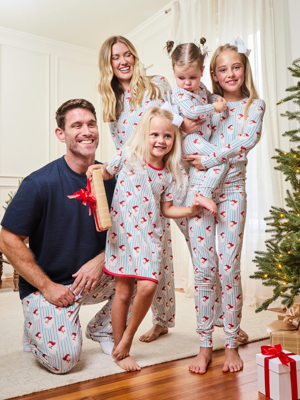 A family of five wearing matching Christmas-themed pajamas poses together in a cozy living room setting with a decorated Christmas tree and wrapped gifts.