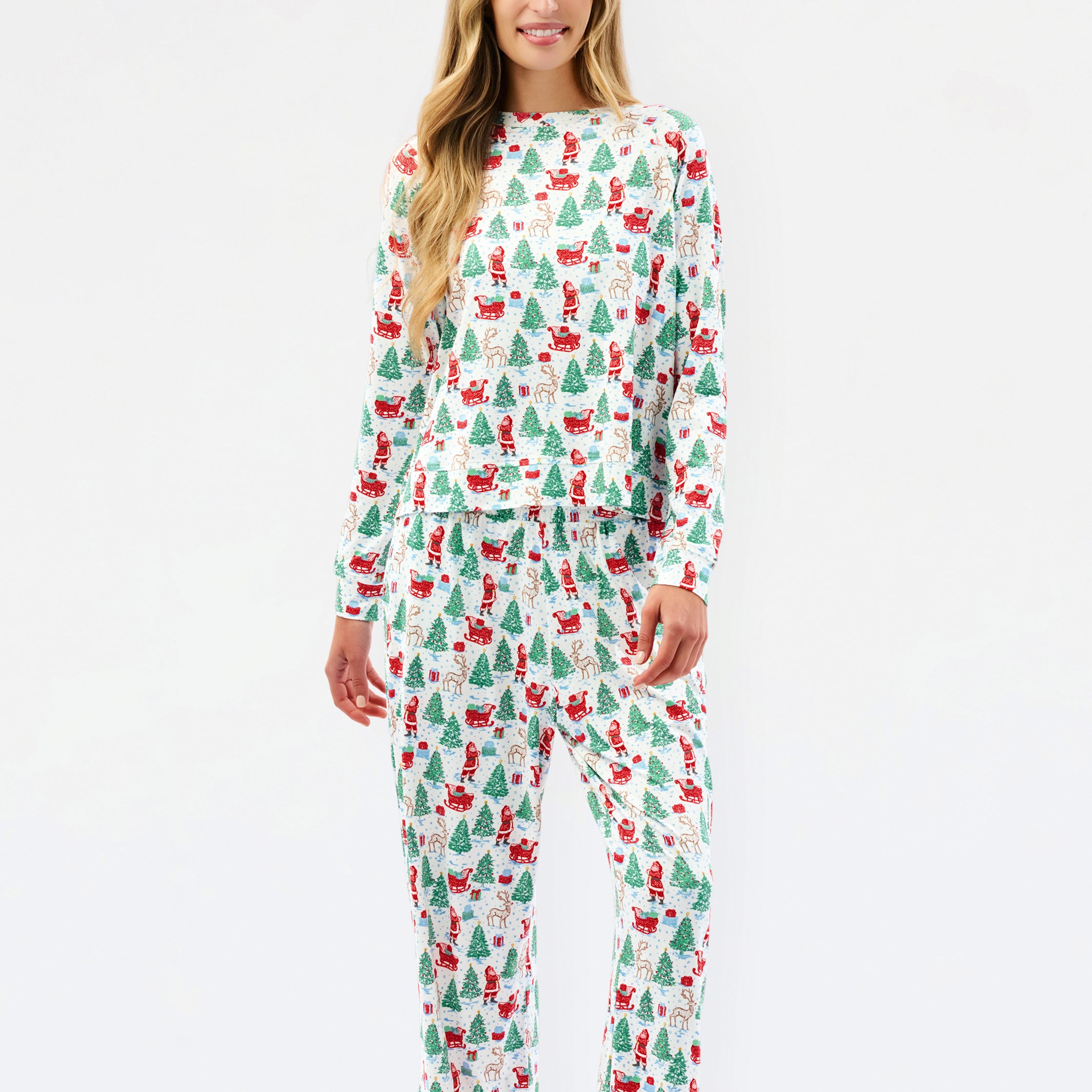 A woman modeling a matching long-sleeve top and pants pajama set with a festive Christmas print of Santa Claus, sleighs, and green trees on a white background.