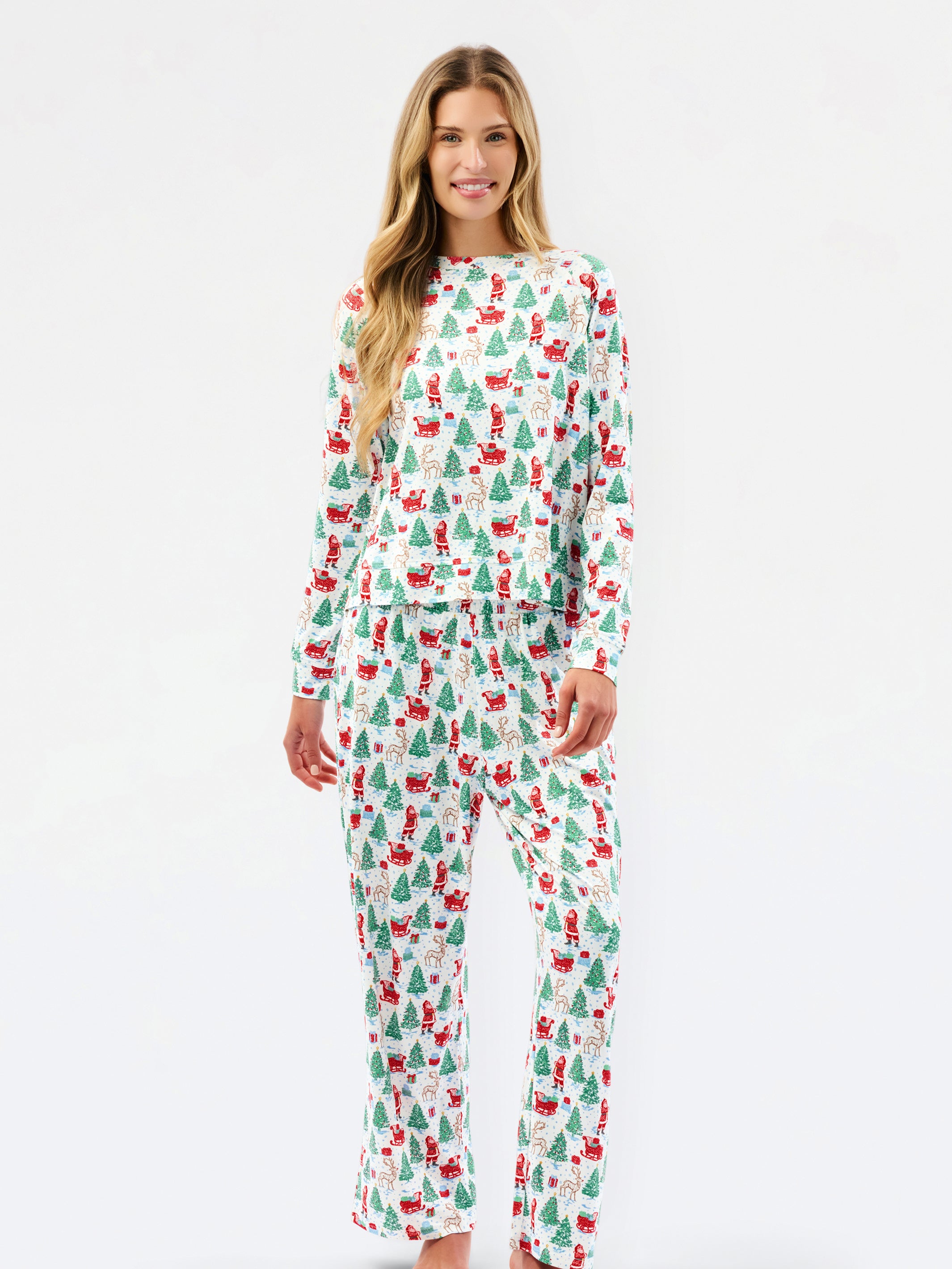 A woman modeling a matching long-sleeve top and pants pajama set with a festive Christmas print of Santa Claus, sleighs, and green trees on a white background.