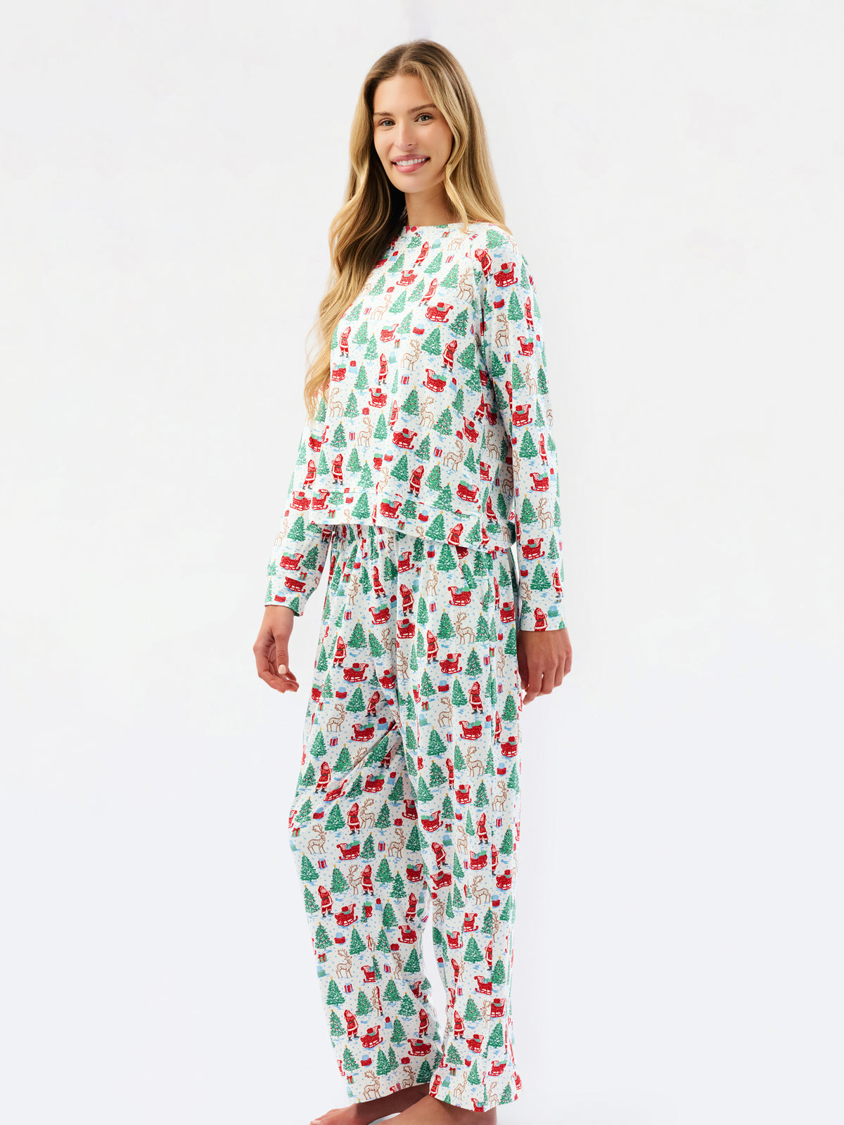 A woman modeling a matching long-sleeve pajama set with a festive Christmas print of Santa Claus, sleighs, and green trees on a white background, photographed against a seamless white studio backdrop.