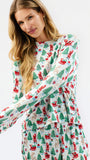 A woman wearing a long-sleeve holiday-themed dress with an all-over print of Santa Claus, Christmas trees, and sleighs on a white background, photographed against a clean white studio backdrop.