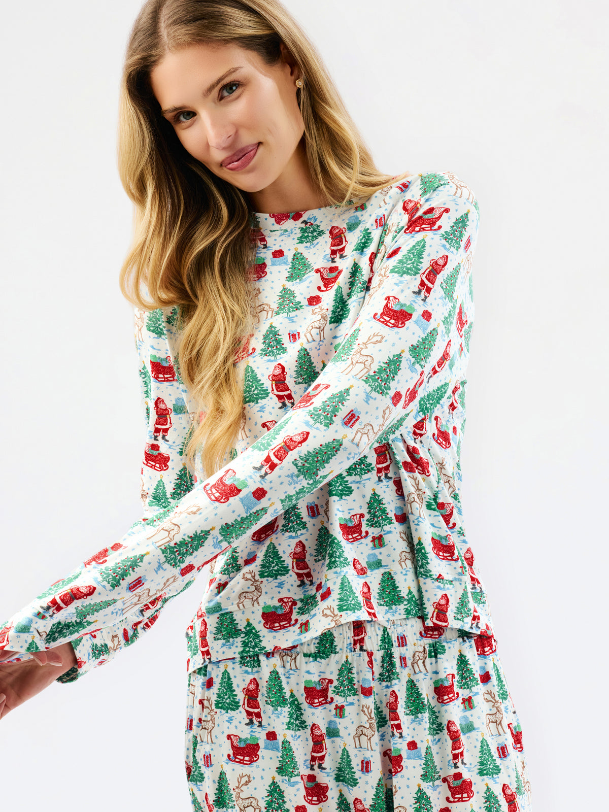 A woman wearing a long-sleeve holiday-themed dress with an all-over print of Santa Claus, Christmas trees, and sleighs on a white background, photographed against a clean white studio backdrop.