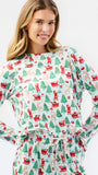 A woman wearing a long-sleeve holiday-themed pajama top with a matching drawstring waist, featuring an all-over print of Santa Claus, reindeer, and Christmas trees on a white background.