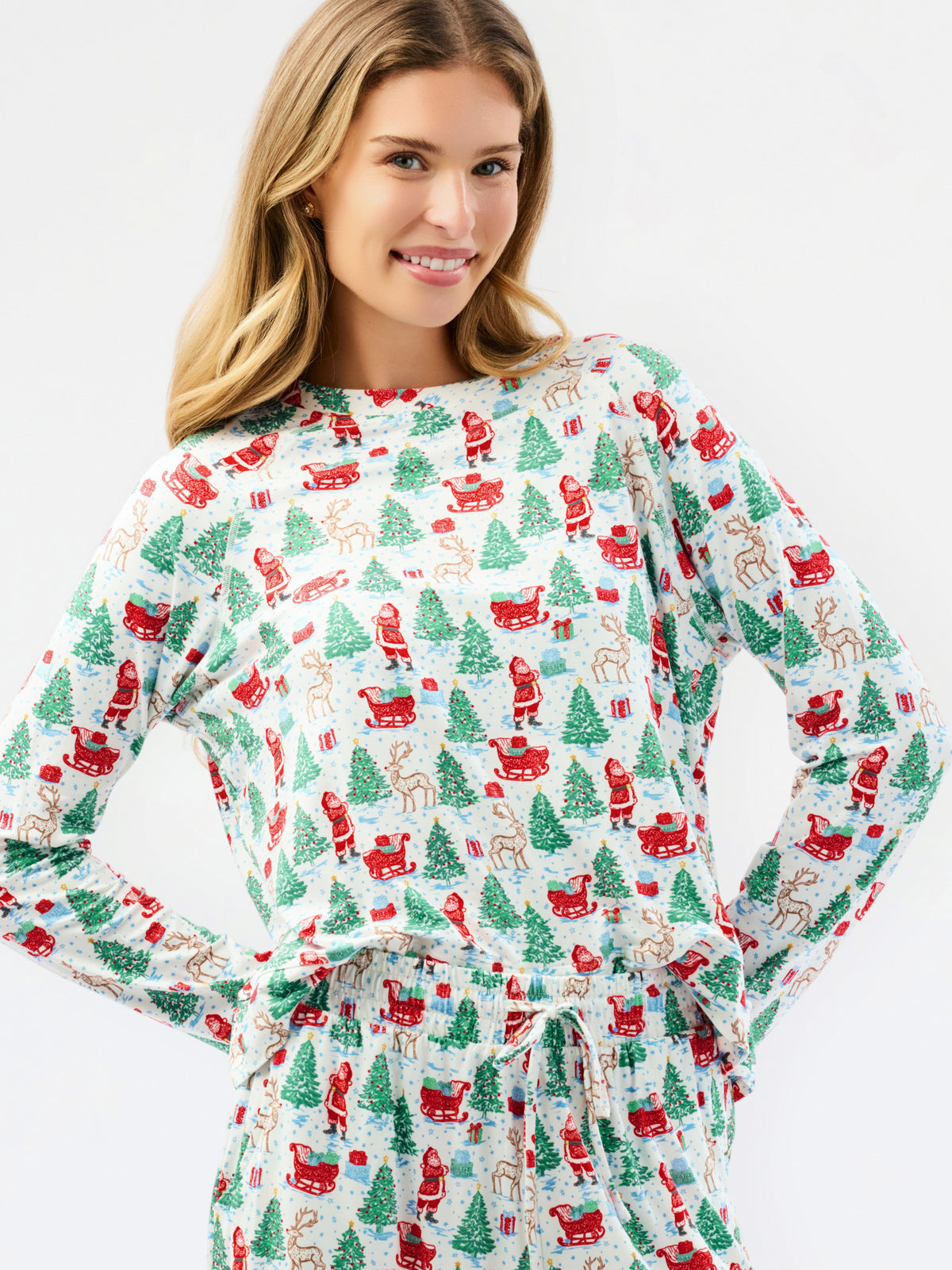 A woman wearing a long-sleeve holiday-themed pajama top with a matching drawstring waist, featuring an all-over print of Santa Claus, reindeer, and Christmas trees on a white background.