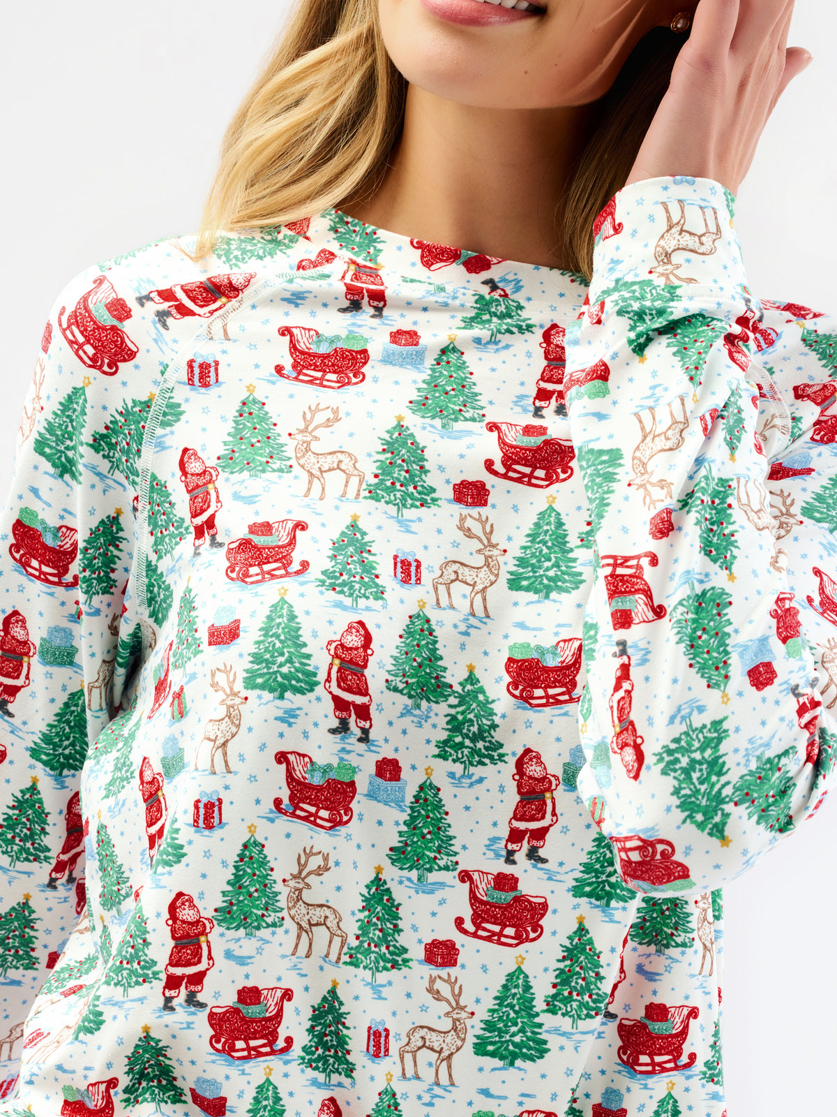 A close-up of a woman wearing a long-sleeve crewneck top with a festive Christmas print featuring Santa Claus, reindeer, sledding, and Christmas trees on a white background.