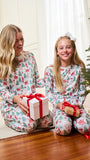 A woman and a young girl sitting on a wooden floor in a living room, both wearing matching long-sleeve pajamas with a festive Christmas tree and Santa Claus pattern. The girl holds a wrapped gift with a red ribbon, and a Christmas tree is visible in the background.