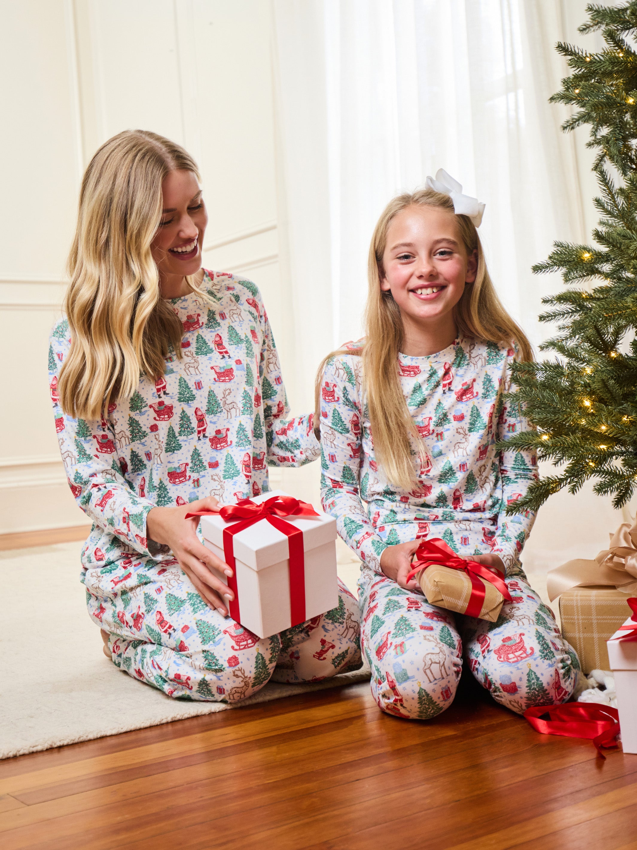 A woman and a young girl sitting on a wooden floor in a living room, both wearing matching long-sleeve pajamas with a festive Christmas tree and Santa Claus pattern. The girl holds a wrapped gift with a red ribbon, and a Christmas tree is visible in the background.